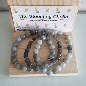 May include: Three beaded bracelets with grey and silver beads are displayed on a wooden block. The Blooming Cholla logo and contact information are visible on a card in the background. The bracelets are handmade jewellery.
