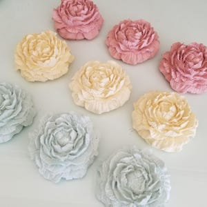 May include: Nine artificial peony flowers are arranged on a white surface. The flowers are in pastel shades of pink, yellow, and blue-grey. The flowers have detailed petal designs. The image is taken from above, showing the flowers in a scattered pattern.