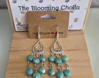 May include: A pair of handmade earrings with a silver teardrop design and turquoise beads. The earrings are displayed on a wooden surface with the text "The Blooming Cholla" and "Handmade with love".