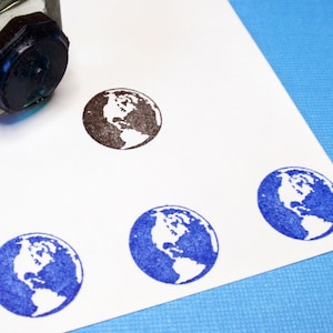 Puede incluir: Un frasco de tinta de vidrio transparente con una almohadilla de sello negra se coloca cerca de varias impresiones de la Tierra. Los sellos de la Tierra son en tinta marrón y azul sobre una superficie blanca. La imagen está sobre un fondo azul claro.