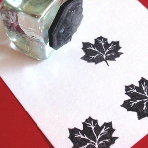 May include: A white card with four black maple leaf stamps. The stamps are arranged in a grid pattern. A clear glass ink bottle with a black rubber stamp is in the upper left corner of the image.