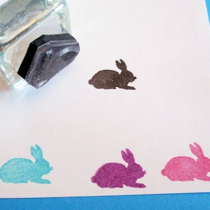 May include: Four rubber stamps of bunnies in different colors. The stamps are on a white surface. The bunnies are black, turquoise, purple, and pink.