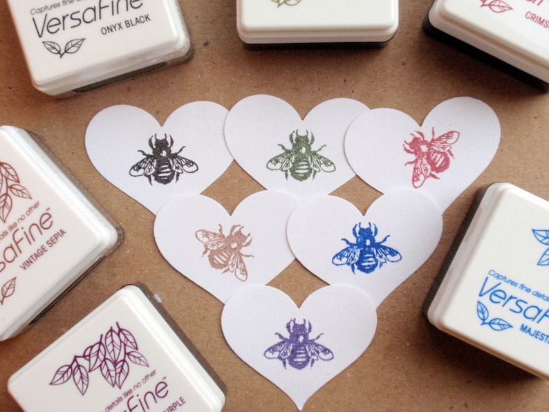 May include: A set of six heart-shaped cards featuring bee illustrations in various colours, including black, green, red, blue, and purple. The cards are arranged in a heart shape on a brown surface. The cards are surrounded by ink pads with the brand name "Versafine" and colour names such as "Onyx Black", "Vintage Sepia", and "Majesty".