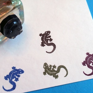 May include: Four rubber stamps of lizards in different colors. The stamps are on a white surface. The lizards are in various poses, with their tails curled up.