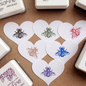 May include: A set of six heart-shaped cards with bee illustrations in various colors, including black, green, red, blue, and purple. The cards are arranged in a heart shape on a brown surface. The cards are surrounded by ink pads with the brand name "Versafine" and color names like "Onyx Black", "Vintage Sepia", and "Majesty".