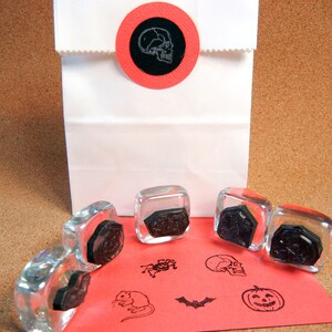 K&ouml;nnte beinhalten: Eine wei&szlig;e Papiert&uuml;te, verschlossen mit einem roten runden Aufkleber mit einem Totenkopf-Design. Klare Glasstempel mit schwarzen Halloween-Motiven, darunter ein Totenkopf, eine Spinne, eine Fledermaus, eine Maus und ein K&uuml;rbis, sind auf rotem Papier angeordnet.