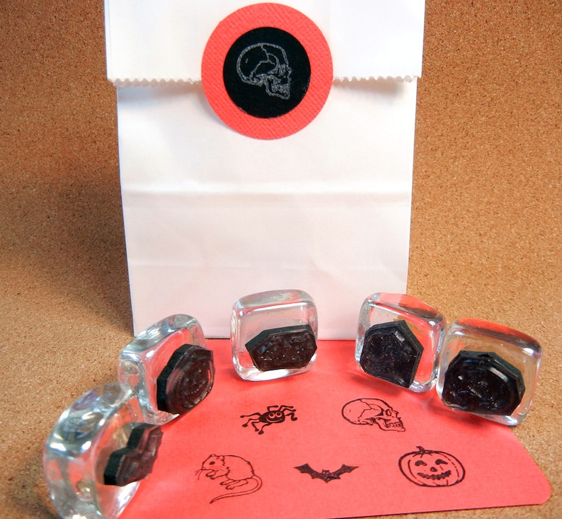 K&ouml;nnte beinhalten: Eine wei&szlig;e Papiert&uuml;te, verschlossen mit einem roten runden Aufkleber mit einem Totenkopf-Design. Klare Glasstempel mit schwarzen Halloween-Motiven, darunter ein Totenkopf, eine Spinne, eine Fledermaus, eine Maus und ein K&uuml;rbis, sind auf rotem Papier angeordnet.
