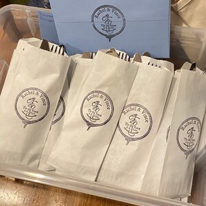 May include: White paper bags with cutlery inside, arranged in a clear plastic container. Each bag is printed with a circular logo that reads "Rachel & Vince Tie the Knot 8.8.2020". A blue envelope with a matching logo is visible.