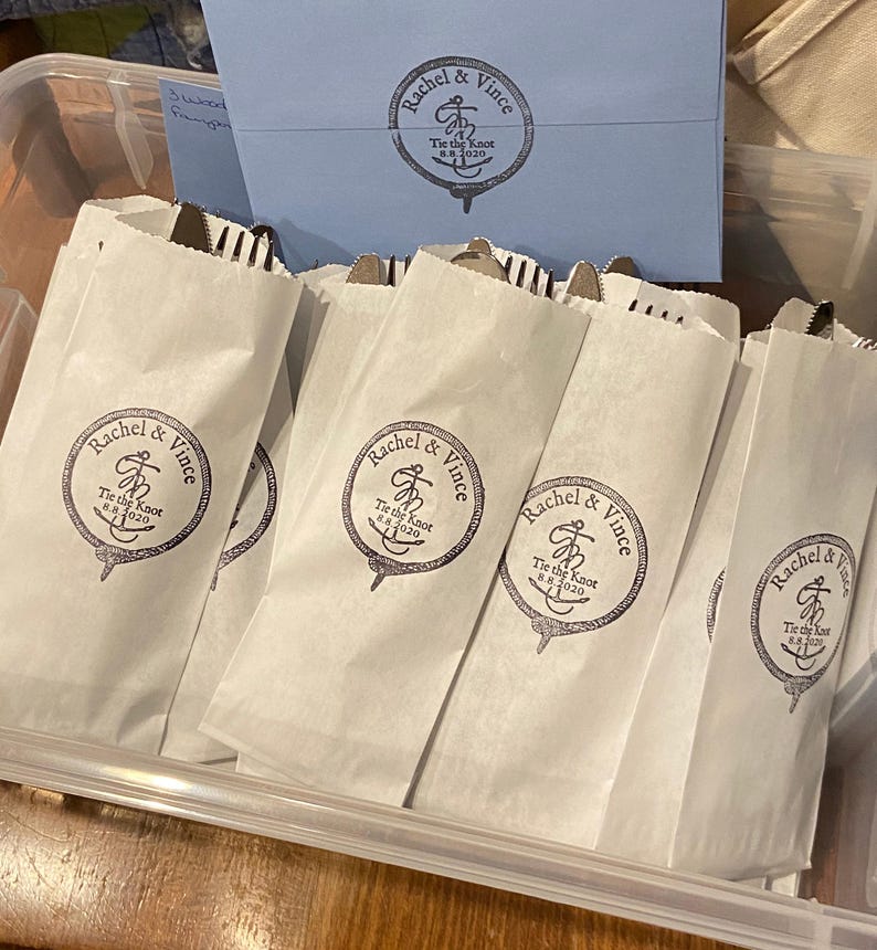 May include: White paper bags with cutlery inside, arranged in a clear plastic container. Each bag is printed with a circular logo that reads "Rachel & Vince Tie the Knot 8.8.2020". A blue envelope with a matching logo is visible.