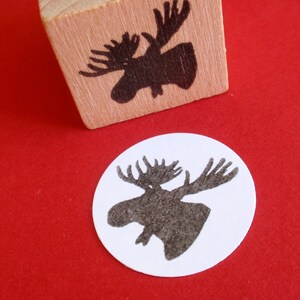 May include: A wooden block stamp with a black silhouette of a moose head. The stamp is on a red surface and a white circle with a black moose head silhouette is next to it.