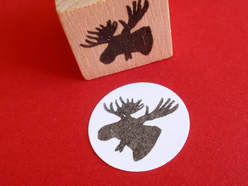 May include: A wooden block stamp with a black silhouette of a moose head. The stamp is on a red surface and a white circle with a black moose head silhouette is next to it.