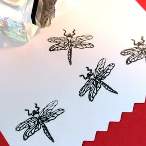 May include: Four black dragonfly rubber stamps on a white surface. The stamps are arranged in a row, with the top stamp slightly offset to the left. The stamps are all the same size and shape, and they feature a detailed dragonfly design.