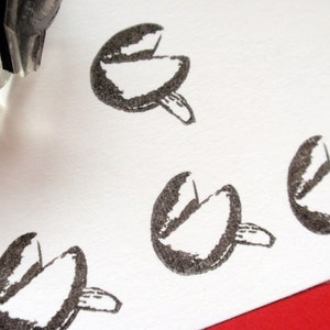May include: Close-up of a stamp creating a pattern of fortune cookies on white paper. The stamp is partially visible in the upper left corner. The fortune cookie impressions are in black.