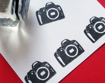 May include: Five black and white rubber stamps of a camera with a lens and a shutter. The stamps are on a white surface.