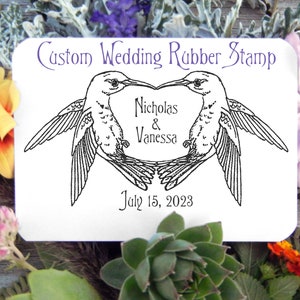 May include: A custom wedding rubber stamp with two hummingbirds facing each other, forming a heart shape. The stamp includes the names "Nicholas & Vanessa" and the date "July 15, 2023" in a decorative font. The design is in black ink on a white background.