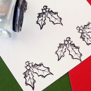 May include: A close-up of a white card with four black and white holly leaf stamps. The stamps are arranged in a diagonal pattern. The card is on a green and red background.