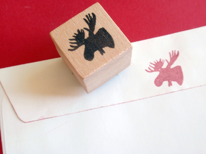 May include: A wooden stamp with a black moose silhouette. The stamp is on a white envelope with a pink moose silhouette.
