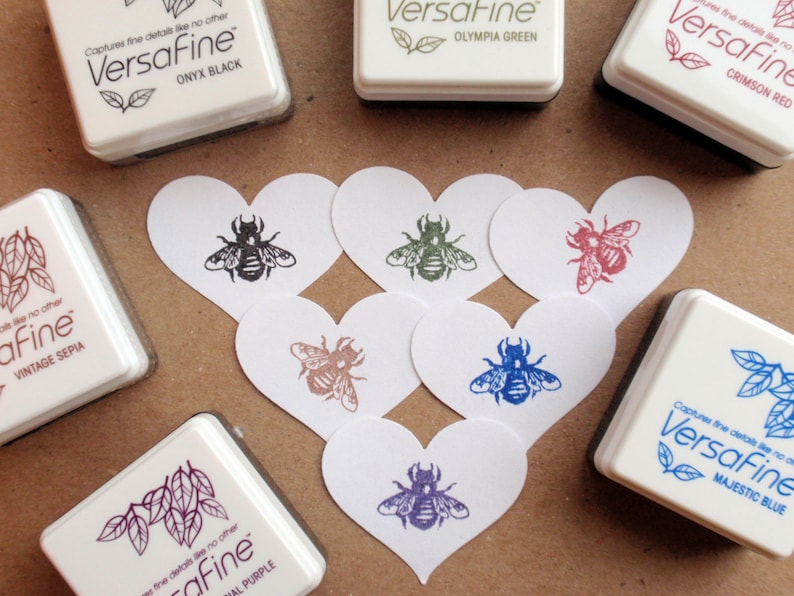May include: A collection of eight ink pads in various colors, each with the text "Versafine" and a color name. The ink pads are arranged around six heart-shaped cards with bee illustrations in different colors.