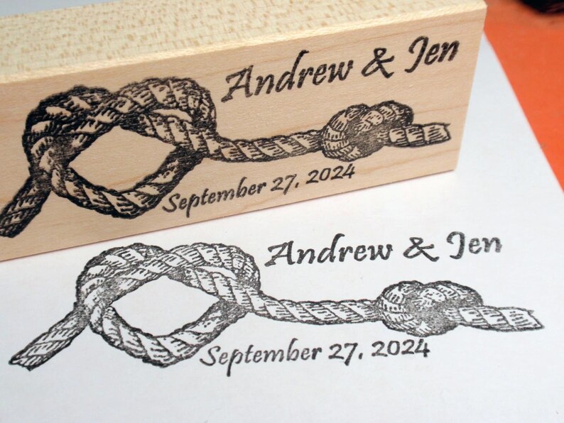 May include: A wooden stamp with a nautical rope knot design. The stamp is engraved with the names "Andrew & Jen" and the date "September 27, 2024". A stamped impression of the design is visible below.