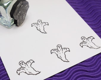 May include: A clear glass stamp with a black ink pad is next to several stamped ghost images on white paper. The ghost stamps are black and have a simple, cartoon-like design. The background is a purple fabric.