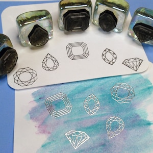 May include: A set of six clear glass stamp pads with silver ink, each featuring a different geometric diamond shape. The stamps are arranged on a white card with a watercolor background.