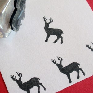 May include: A clear glass stamp with a black deer silhouette design. The stamp has been used to create multiple black deer prints on a white paper surface. The background is a solid red color.