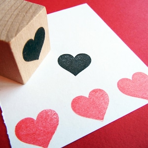 May include: A wooden block with a black heart design is used to stamp four hearts onto a white card. The hearts are red and black.