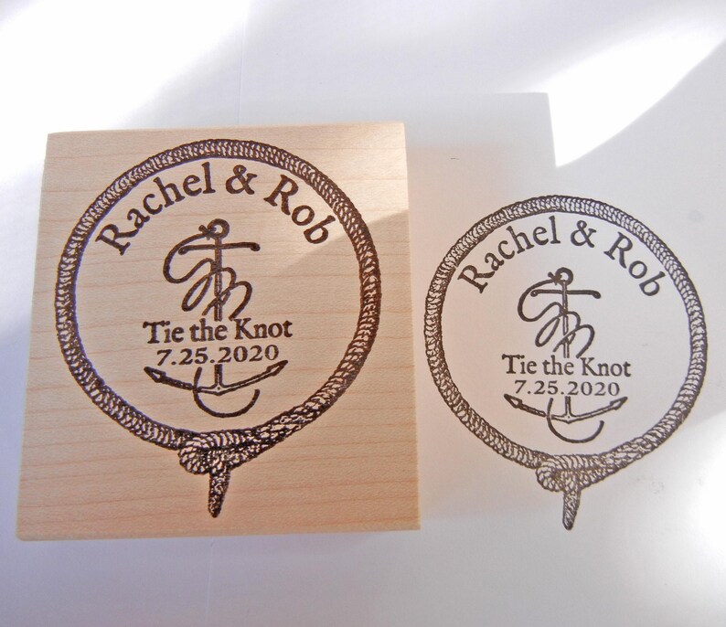 May include: A wooden stamp with a circular design featuring the names "Rachel & Rob", the phrase "Tie the Knot", and the date "7.25.2020". The design includes an anchor and rope border.