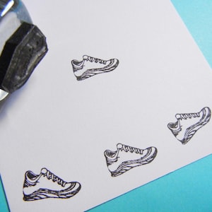 May include: A clear stamp with a black rubber shoe design is shown in the upper left corner. The stamp has been used to create multiple black shoe prints on a white card. The card has rounded corners.