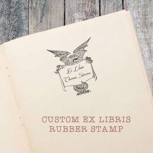 Może przedstawiać: Otwarta strona książki z niestandardowym projektem gumowej pieczątki Ex Libris. Pieczątka przedstawia szczeg&oacute;łową ilustrację skrzydlatej istoty trzymającej transparent z napisem "Ex Libris Thomas Stevens". Poniżej ilustracji tekst "CUSTOM EX LIBRIS RUBBER STAMP" jest wydrukowany w kolorze czerwonobrązowym.