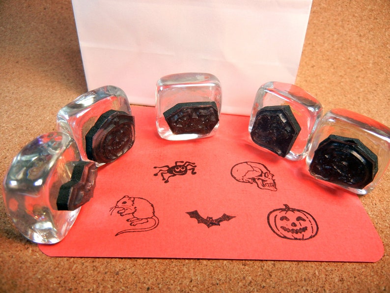 May include: Five clear glass stamp blocks with black rubber stamps inside. The stamps feature Halloween themed designs including a skull, a pumpkin, a bat, a spider, a mouse, and a geometric design.