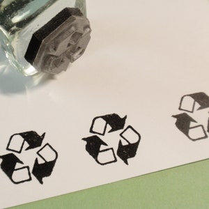 May include: Three black rubber stamps of the universal recycling symbol on a white surface. The stamps are made of black rubber and have a raised design. The symbol is a circle with an arrow pointing to the right, and the arrows are pointing in a clockwise direction.