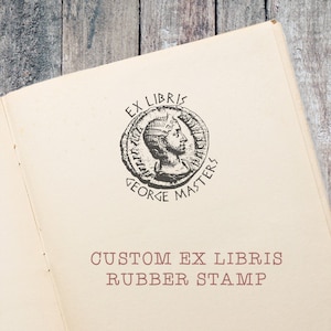 May include: Open book with a cream-colored page featuring a circular stamp design. The stamp includes the words "EX LIBRIS" and "GEORGE MASTERS" around a profile image of a person. Below the stamp, the text reads "CUSTOM EX LIBRIS RUBBER STAMP."