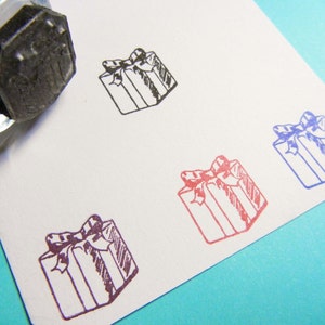 May include: A clear stamp with a black ink pad is positioned near stamped gift box designs. The gift boxes are in black, red, and blue ink on a white card. The gift boxes have a bow on top.