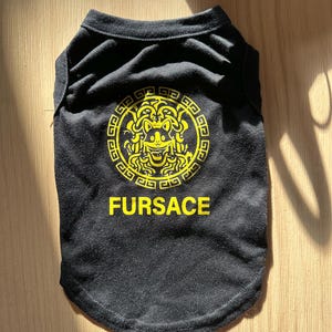 K&ouml;nnte beinhalten: Schwarzes Hunde-T-Shirt mit einem gelben, von Medusa inspirierten Grafikdesign und dem Wort "FURSACE" in Gelb. Das Shirt besteht aus einem weichen Material und ist f&uuml;r Haustiere konzipiert.