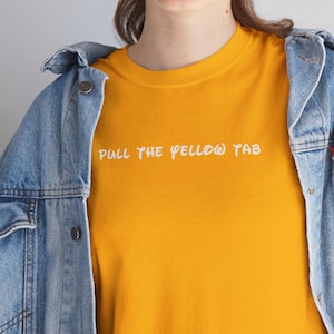 Puede incluir: Camiseta de color amarillo mostaza con el texto blanco "PULL THE YELLOW TAB". La camiseta se combina con una chaqueta vaquera azul claro. La camiseta es una prenda informal.