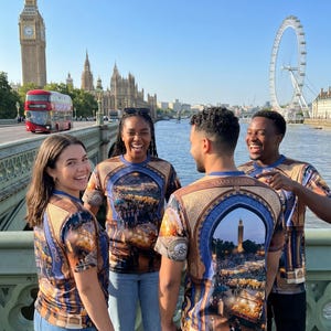 Könnte beinhalten: Vier Personen tragen T-Shirts mit einer detaillierten Grafik einer Stadtszene, möglicherweise Marrakesch, mit einem blau-braunen Design. Die Shirts tragen das Wort "Marrakech" auf dem Rücken. Der Hintergrund zeigt Londoner Wahrzeichen.