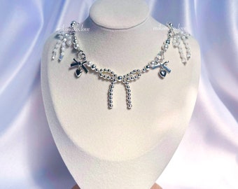 May include: A silver necklace with a delicate design, featuring small bows and heart-shaped charms. The necklace is adorned with small beads and dangling crystal accents. The necklace is displayed on a white bust against a soft, light blue background.