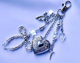 May include: Silver-toned keychain with a heart-shaped locket, adorned with charms including a bow, cross, star, and beads. The locket has an engraved floral design. The keychain is attached to a silver clasp.