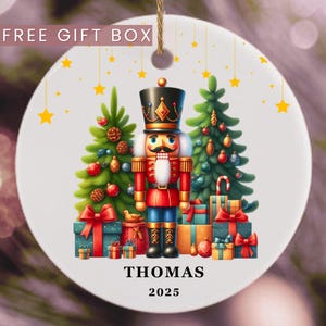 May include: A white ceramic Christmas ornament featuring a colorful illustration of a nutcracker soldier, Christmas trees, and wrapped gifts. The ornament has the name "THOMAS" and the year "2025" printed on it. The words "FREE GIFT BOX" are at the top.