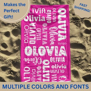 May include: A bright pink beach towel with the name "Olivia" in white, multiple fonts. The towel has the text "Multiple Colors and Fonts". The background is sand.