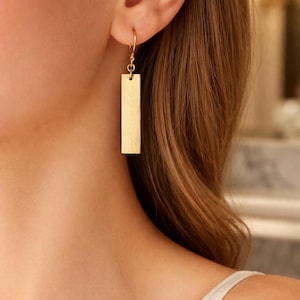 May include: A gold-colored rectangular earring hangs from a delicate hook. The earring is a simple, elongated rectangle with a textured surface. The image shows the earring in detail, highlighting its design and color.