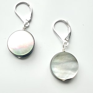 May include: A pair of drop earrings featuring circular, iridescent mother-of-pearl discs suspended from silver-toned leverback hooks. The discs display a smooth, polished surface with subtle variations in white, gray, and soft pink tones.