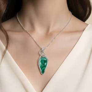 May include: A teardrop-shaped malachite pendant featuring deep green swirling patterns, encased in a silver wire-wrapped setting. The pendant hangs from a delicate silver chain, resting against a plain cream-colored fabric background.