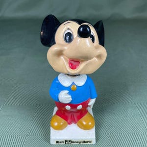 May include: A vintage Mickey Mouse bobblehead figurine. Mickey is wearing a blue shirt, red shorts, and yellow shoes. The base is white with the words "Walt Disney World" printed on it. The figurine has black ears and a smiling face.