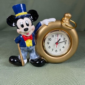 May include: A ceramic Mickey Mouse figurine in a blue suit and top hat, holding a cane, stands next to a gold-colored Seiko quartz clock. The clock face is white with black numerals and hands. The clock is in the shape of a pocket watch.