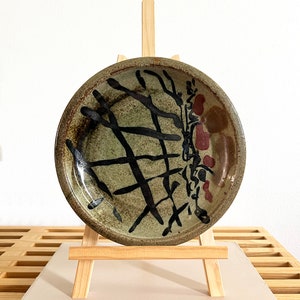 May include: A ceramic plate with a brown, green, and black glaze. The plate has a geometric design with black lines and red accents. The plate is displayed on a wooden easel.