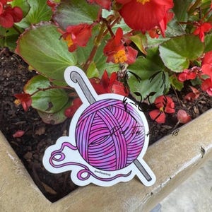 May include: A sticker featuring a pink and purple yarn ball with a crochet hook. The sticker is set against a background of red flowers and green leaves. The text "redfoxarts48" is visible.