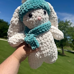 May include: A handmade, white crochet bunny wearing a teal hat and scarf. The bunny has black eyes and a pink nose. The plush toy is soft and fluffy, perfect for a child's gift or nursery decor.