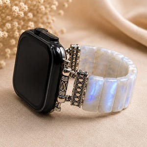 Könnte beinhalten: Eine schwarze Smartwatch mit einem dekorativen silberfarbenen Metallanschluss, befestigt an einem dehnbaren Armband aus rechteckigen, schillernden Mondsteinperlen, die im Licht einen sanften blauen Schimmer zeigen.